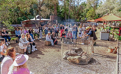 Large group of people gathered around a campfire, listening to a demonstration.