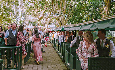 Guests in formal attire board a covered train car, traversing a paved walkway lined with trees.