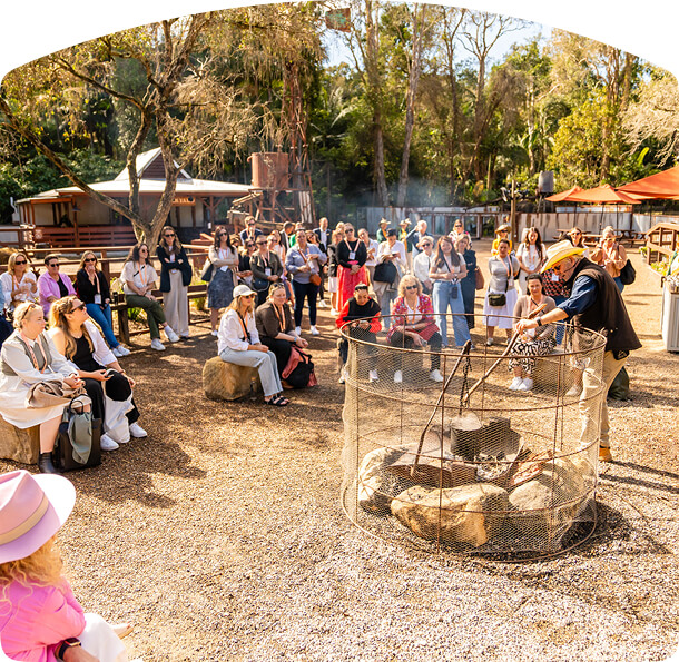 A man in a yellow hat demonstrates cooking over an open fire to a seated audience outdoors.