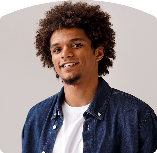 Person with curly hair wearing a dark blue denim shirt over a white t-shirt, smiling slightly while looking at the camera.