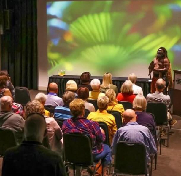 Indoor seminar with a speaker in traditional attire presenting near a podium labeled WILD, vibrant nature visuals on screen, and audience attentively seated