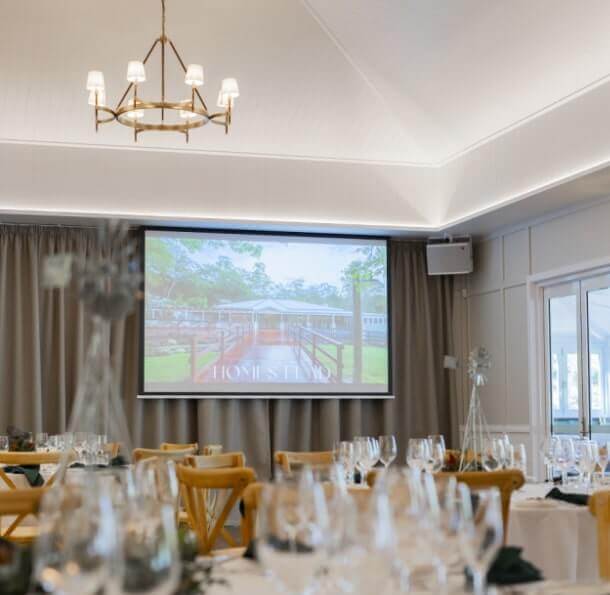Sophisticated event space with vaulted ceiling, chandelier, projection screen showing HOMESTEAD, and tables set with wine glasses and greenery centerpieces