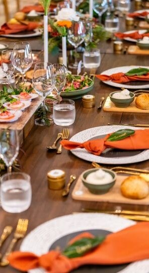 Formal dining table arranged with black plates orange napkins topped with green leaves gold cutlery bread on wooden trays and floral centerpieces