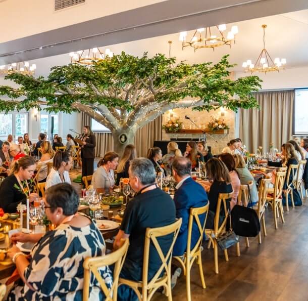 Large indoor dining event with guests seated at long wooden tables beneath an artificial tree centerpiece, chandeliers, and floral arrangements in a sophisticated setting