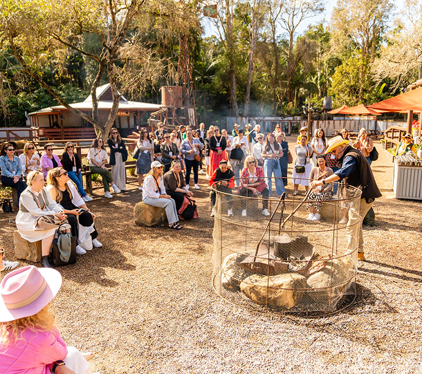 Large group of people gathered around a campfire, listening to a demonstration.