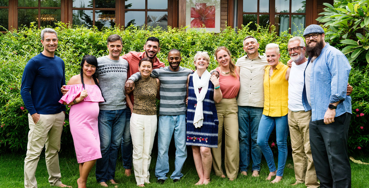 Group of twelve adults standing closely together outdoors in front of a green hedge and building with large windows.