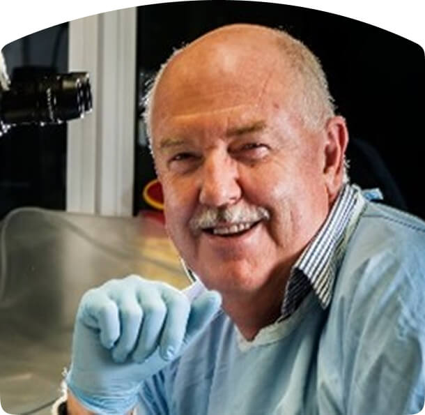 Older individual in a light blue medical gown and blue gloves, smiling while seated in a laboratory or clinical environment.