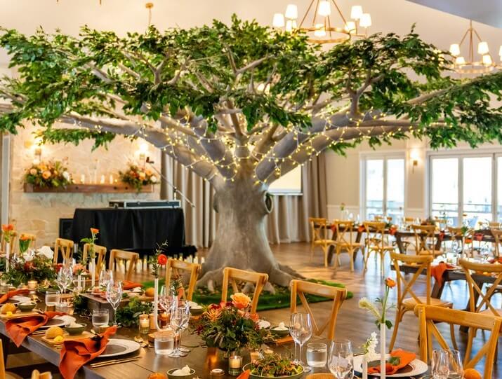 Decorated event space featuring a large artificial tree wrapped in string lights surrounded by wooden tables with orange napkins floral centerpieces and candles under chandeliers and natural light
