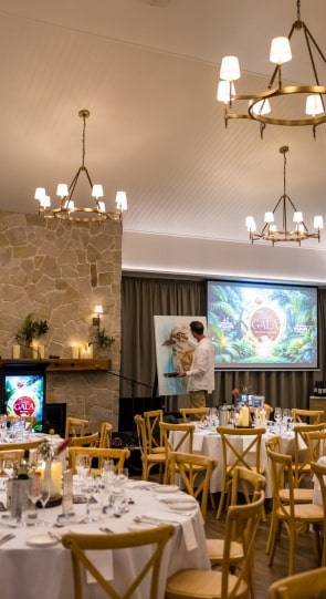 A corporate event setting with elegantly set tables, a man in white attire presenting near a screen displaying a gala logo.