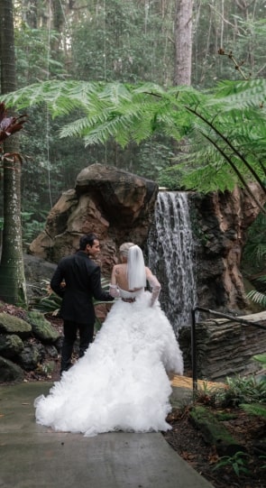Bride and groom standing together, gazing at a waterfall surrounded by lush greenery in a peaceful forest setting.