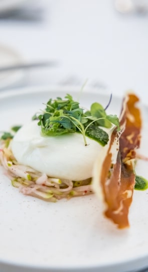 A plated dish featuring a poached egg topped with microgreens and crispy prosciutto, served on a bed of shredded vegetables.