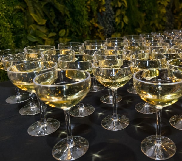 A table filled with numerous champagne coupes filled with sparkling liquid, set against a lush green backdrop.