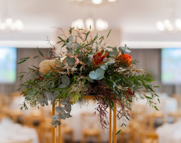A floral centerpiece with various greenery and orange flowers, displayed on a table in a decorated event space.