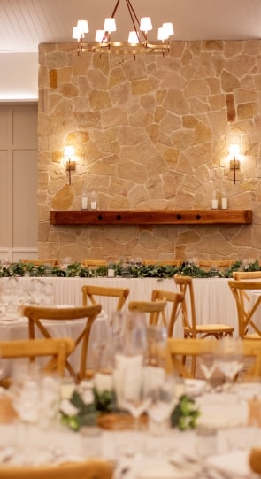 Elegant wedding reception setup featuring wooden tables, stone wall with candles, and greenery accents.
