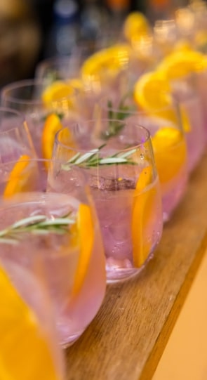 Rows of glasses filled with pink beverages garnished with orange slices and rosemary, displayed on a wooden surface.