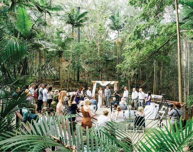 Outdoor wedding ceremony in forest setting with bride, groom, and guests gathered around floral altar