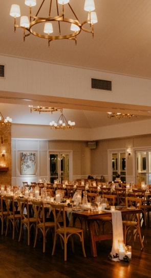 A beautifully set dining area for a wedding, featuring wooden tables, candles, and elegant chandeliers, illuminated softly.
