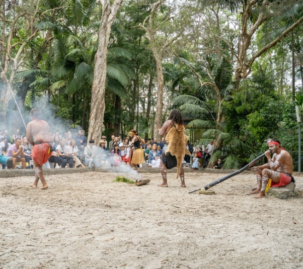 Three performers in traditional attire engage in cultural activities in a forest setting, with an audience watching.