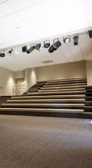View of an empty auditorium with tiered seating and overhead stage lights, designed for presentations and corporate events.