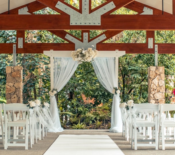 Wedding ceremony setup featuring white chairs, an aisle runner, and a floral arch surrounded by greenery.