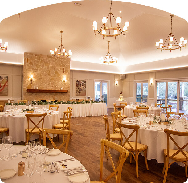 Indoor dining area with round tables covered in white linens, wooden chairs, a long head table with greenery, and a stone wall featuring candles and soft lighting