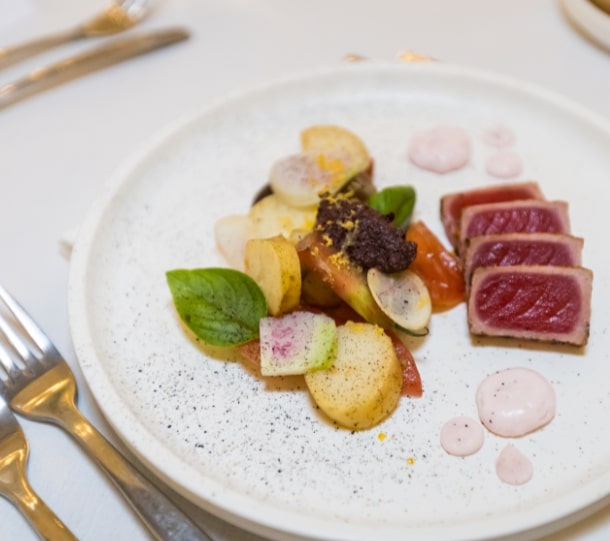 Plate of seared tuna slices alongside colorful vegetables and sauce, displayed on a textured white dish.