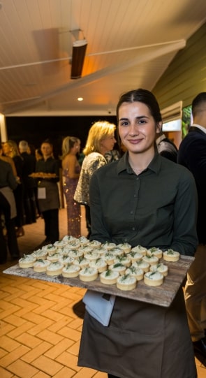A server in a green shirt holds a wooden tray of canapés at a corporate event, with guests mingling in the background.