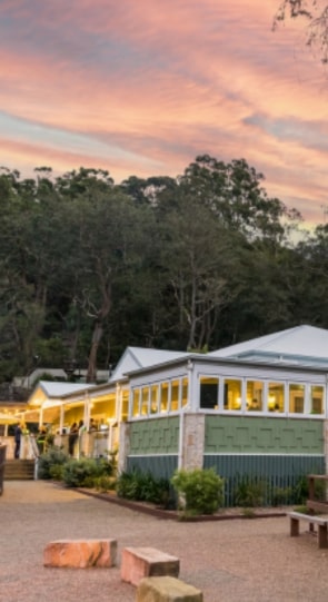 Sunset view of a green-and-white building surrounded by trees, with outdoor seating and a gravel pathway in front.