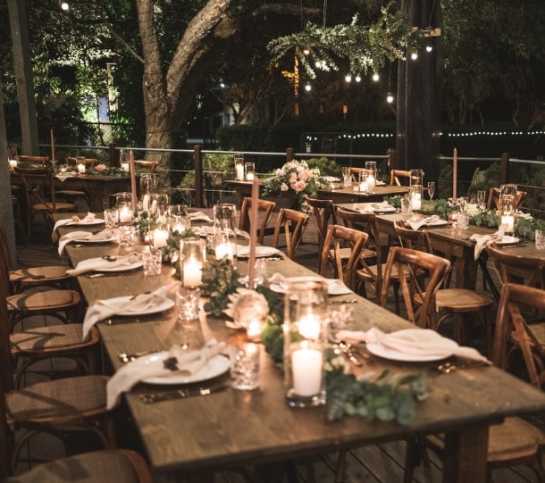 Elegant outdoor dining setup with woodean tables, candles, and floral centerpieces, illuminated by string lights.