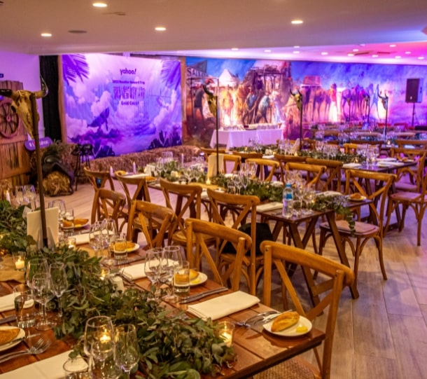Elegant dining setup for a corporate event, featuring wooden tables with greenery, glassware, and a decorative backdrop.