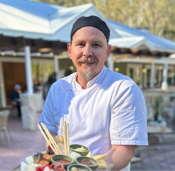 Chef in white uniform and black hat holding a platter of assorted appetizers with dip bowls, standing outdoors.