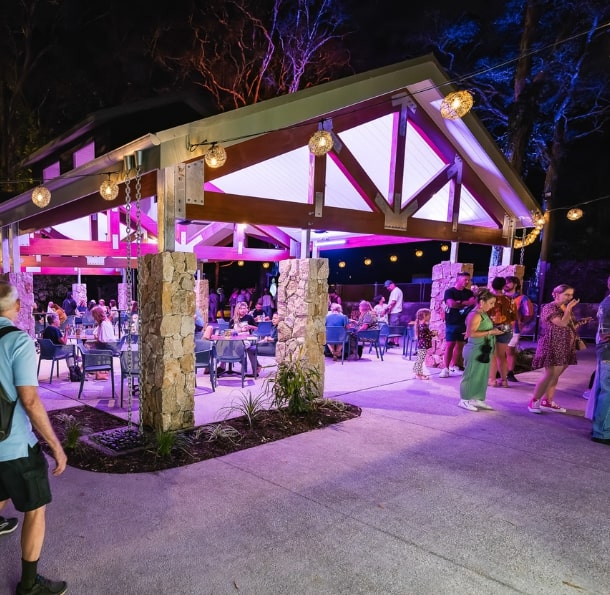 Vibrant outdoor venue lit with colorful lights, featuring guests dining and socializing under a large shelter.
