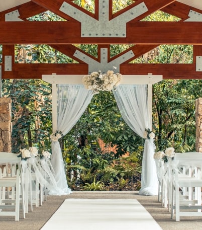 Ceremony setup featuring white draped arch adorned with flowers, surrounded by chairs and lush greenery in a natural setting.