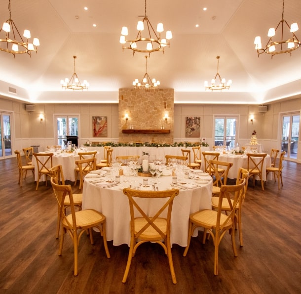 Elegant dining room with round tables set for a gathering, featuring soft lighting, wooden floors, and a stone fireplace.