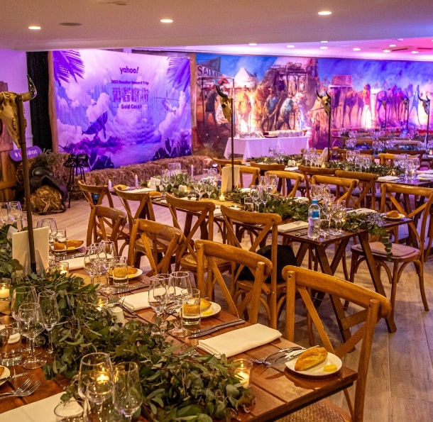 Elegant reception area with wooden tables set for dining, featuring greenery, glassware, and decorative backdrop