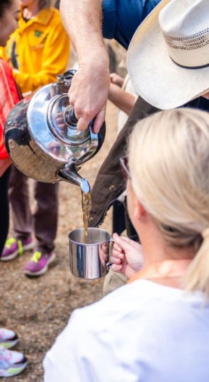 Man in cowboy hat pours tea from a metal kettle into a metal cup held by a blonde woman outdoors