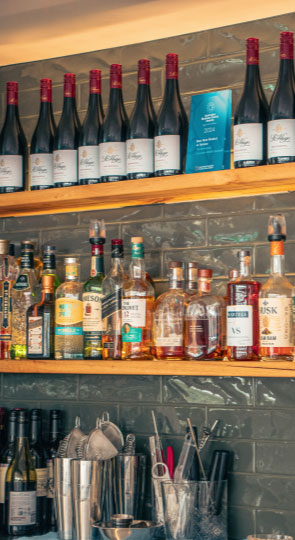 Shelves with rows of red wine bottles and assorted spirits above a bar, cocktail shakers and utensils on the shelf below