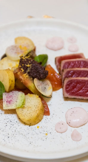 Seared rare tuna with heirloom tomato, baby potatoes, radish slices, pink sauce dots and a dark tapenade on a white plate