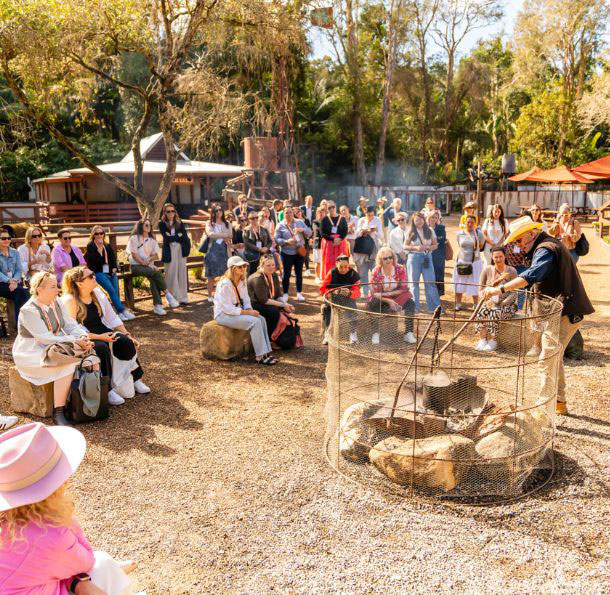 Large group gathered around a campfire at Outback Springs, watching a presenter swing a cooking pot on a sunny day
