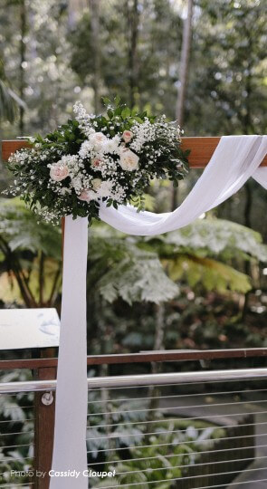 Wedding arch draped in white fabric with bouquet of white and blush-pink roses and greenery against a forest backdrop