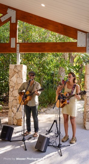 Two musicians singing and playing acoustic guitars beneath a wooden pavilion with stone pillars and sunlit trees