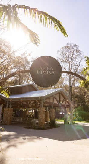 Outdoor pavilion entrance amid trees and palm fronds in bright sun; arched sign reading Astra Lumina