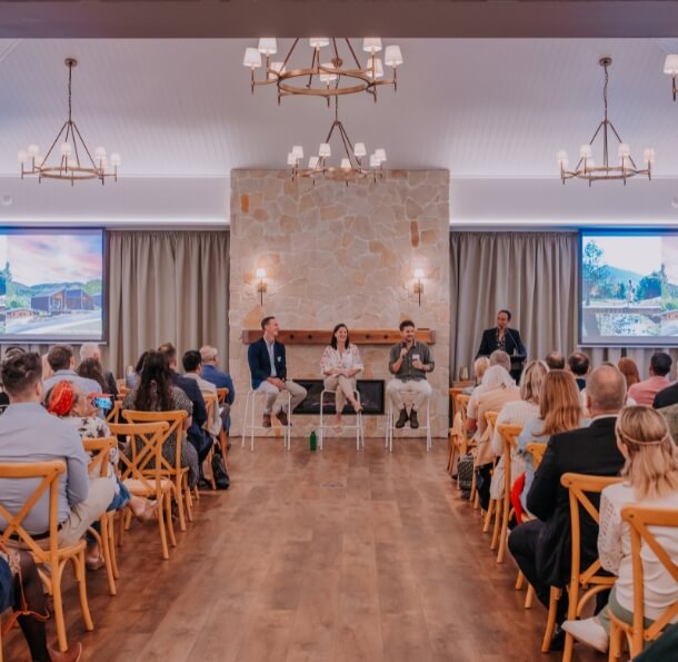 Audience seated in a rustic elegant hall at The Homestead watching three panel speakers on stage with two projection screens