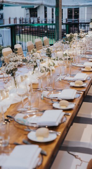 Long wooden banquet table on a deck set for a white wedding with white napkins, rolls, glassware and floral centerpieces.