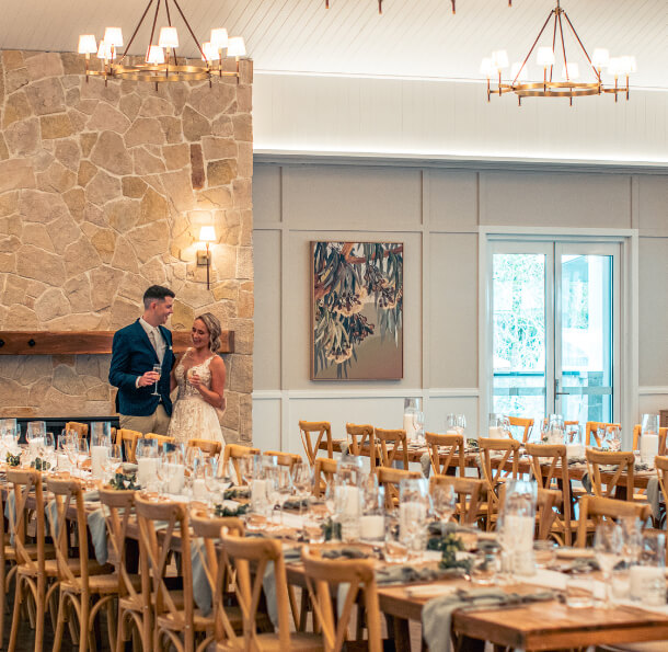 Bride and groom gazing at each other behind a long, elegantly set reception table in a light-filled rustic-modern hall.