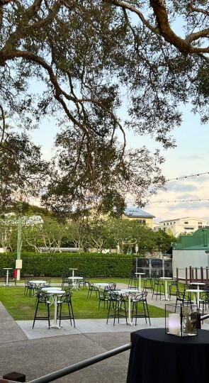 The Village Deck: cocktail tables on grass and concrete beneath a large tree and string lights, hedges in the background.