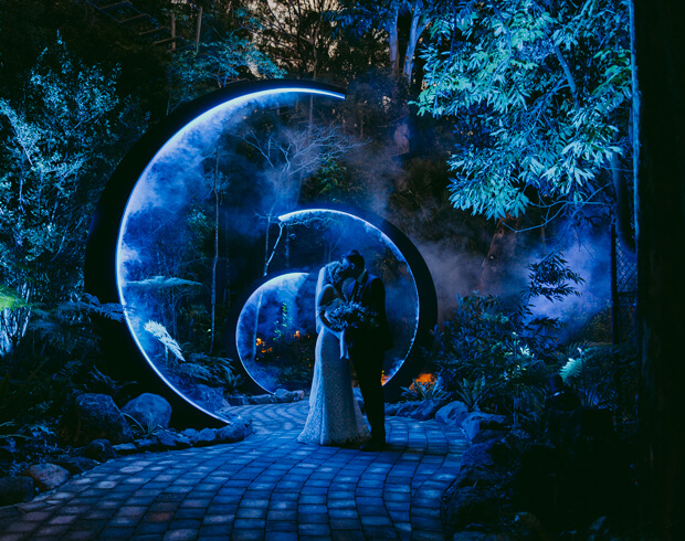 Silhouetted couple kissing under glowing crescent arches in a forest.