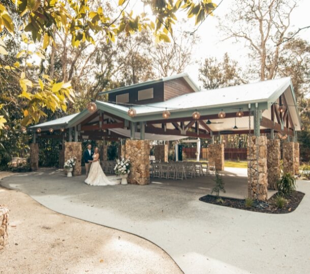 Bride and groom standing under a modern rustic open-air pavilion at a sunlit wooded wedding venue