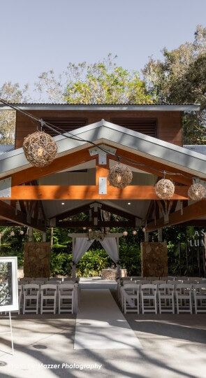 Open-air wooden pavilion set for a wedding: white chairs along an aisle leading to a white-draped altar