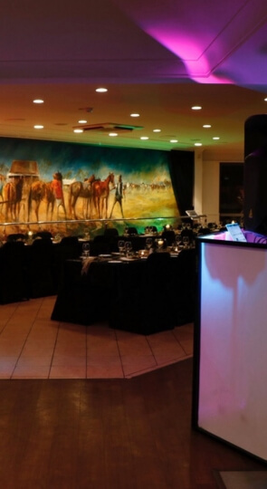 Event room with set dining tables, a lit lectern and speakers, and a large mural of cowboys and horses on the back wall